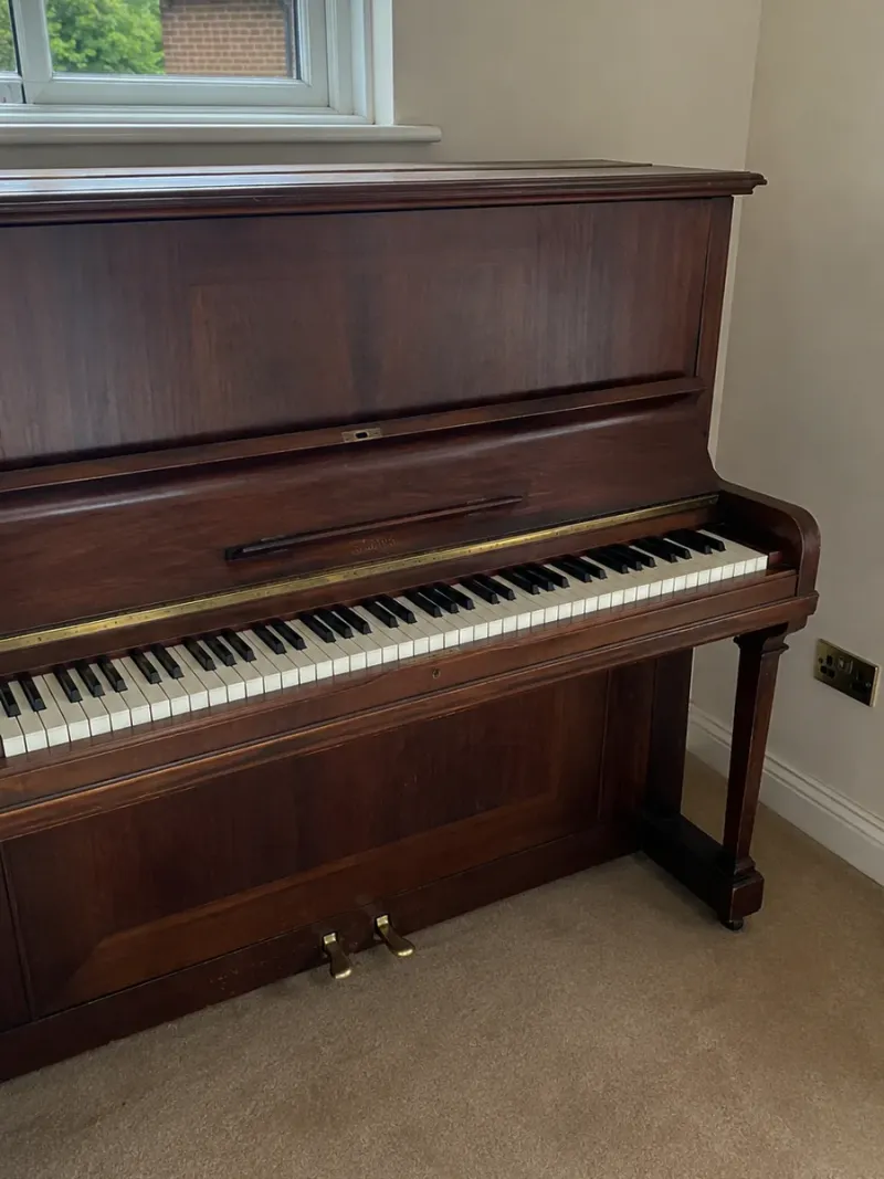 Mahogany upright piano standing in a carpeted room beside a window, the kind of specialist item Bridge Removals moves.